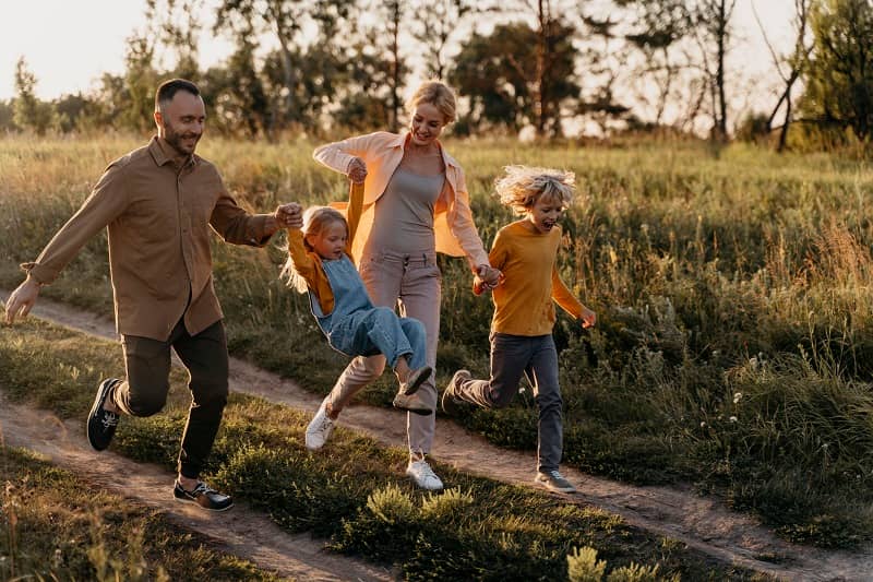 Glückliche Familie mit zwei Kindern rennt lachend über einen Feldweg inmitten grüner Natur bei warmem Sonnenlicht am späten Nachmittag.